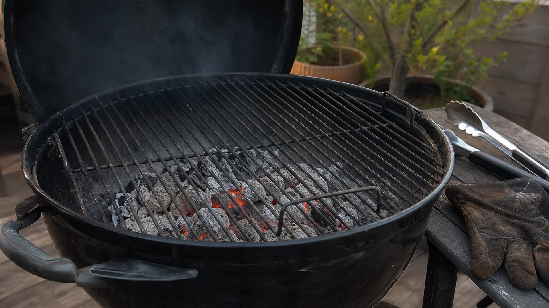 Geöffneter Kugelgrill mit glühender Holzkohle, daneben liegen Grillzange und Grillhandschuh