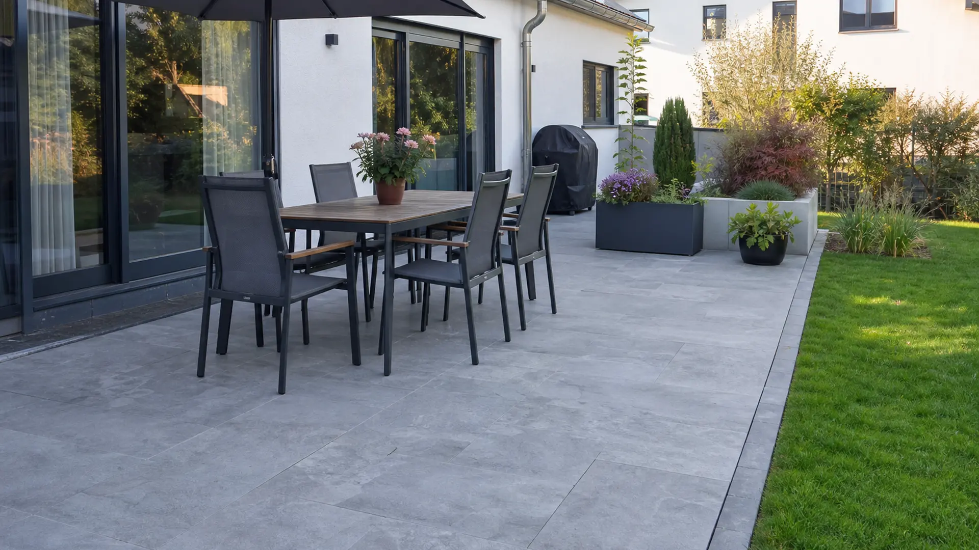 Terrasse mit großformatigen Platten am Haus, darauf ein Esstisch mit Stühlen, dahinter der Rasen