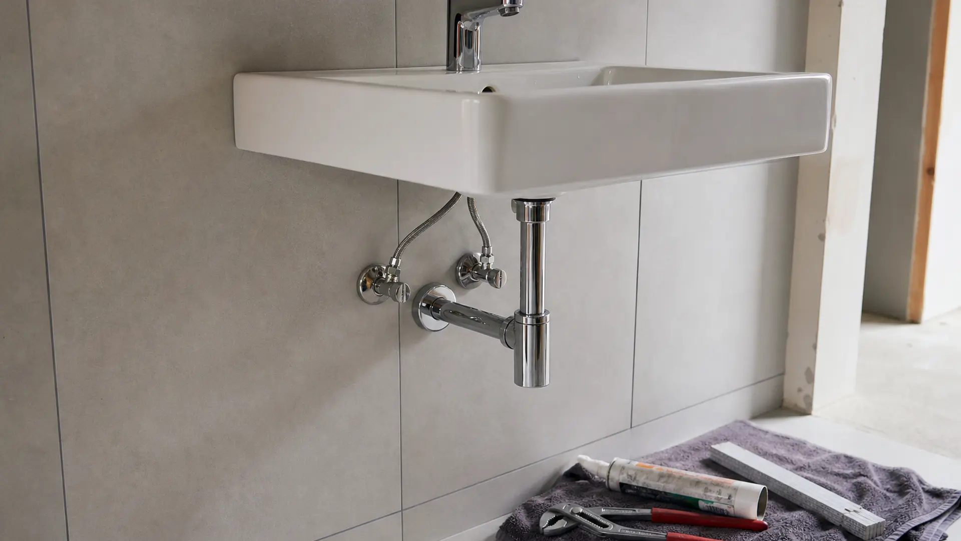 Weißer Waschtisch an einer gefliesten Wand, darunter sichtbar Siphon und Eckventile, Werkzeug liegt auf einem Tuch daneben