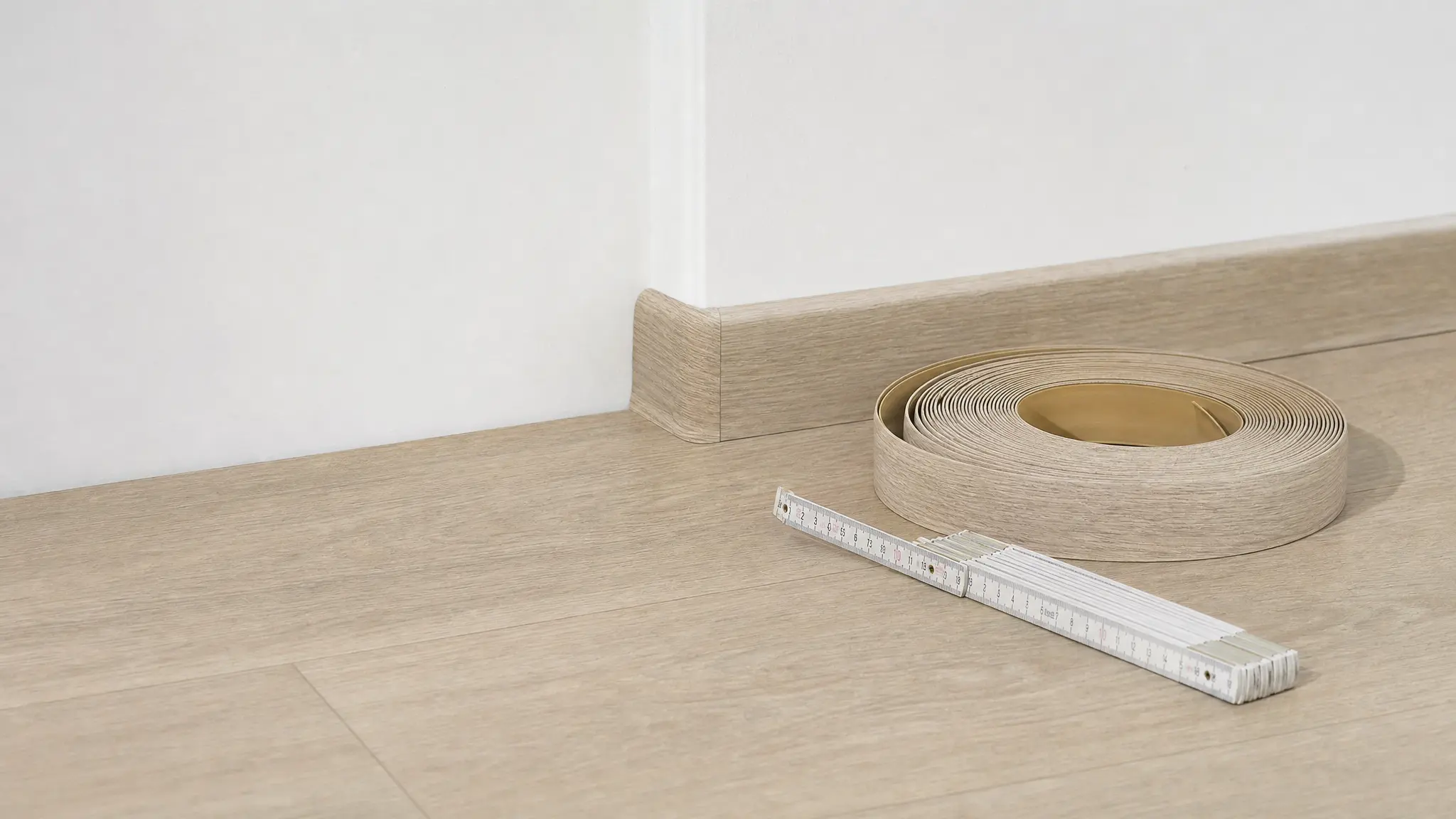 Eine weiße Sockelleiste schließt eine Wand mit hellem Laminat ab, daneben liegen Zollstock und Leistenrolle