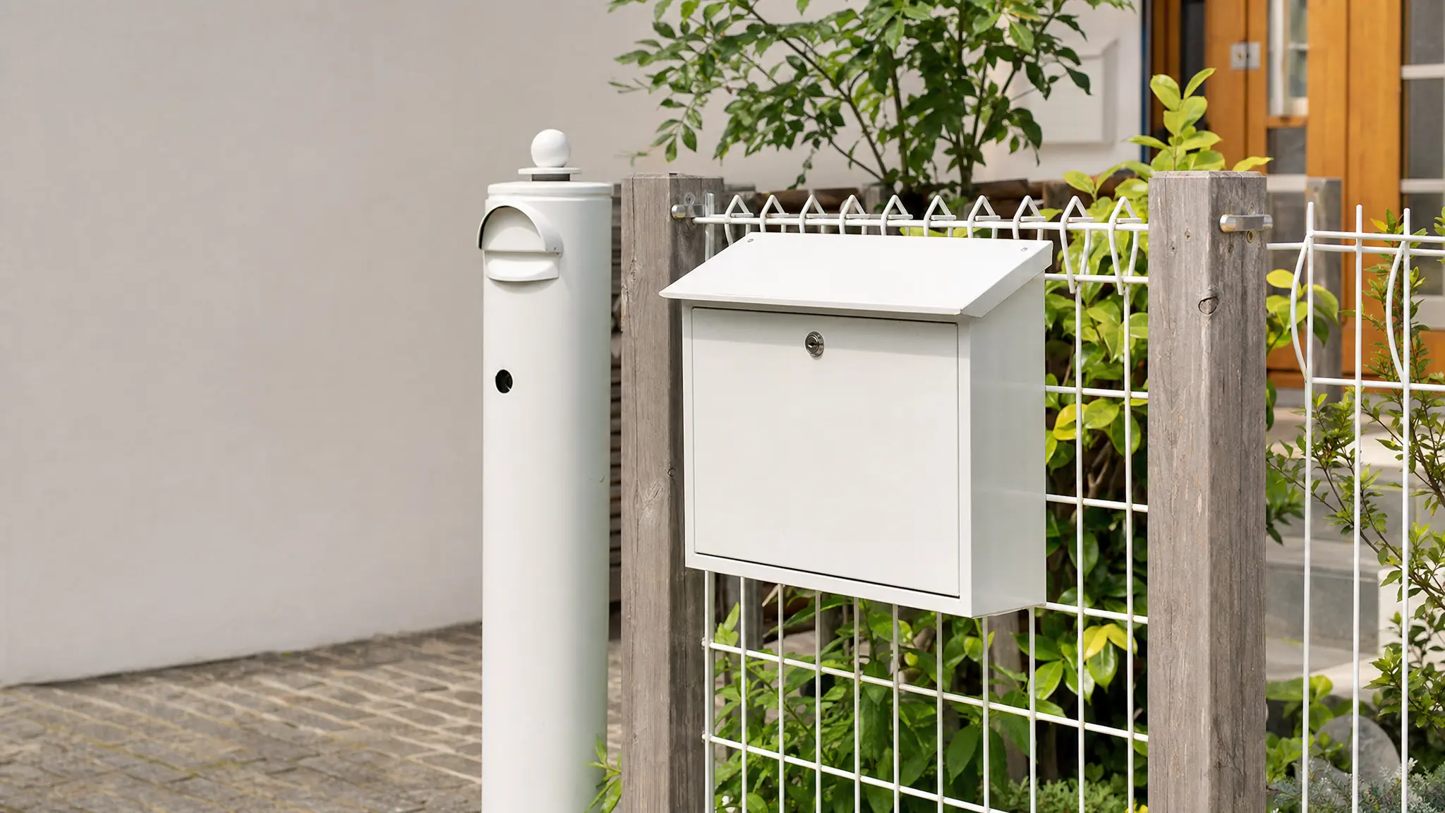 Ein weißer Briefkasten hängt am Zaun neben dem Hauseingang, daneben steht ein Briefkastenpfosten